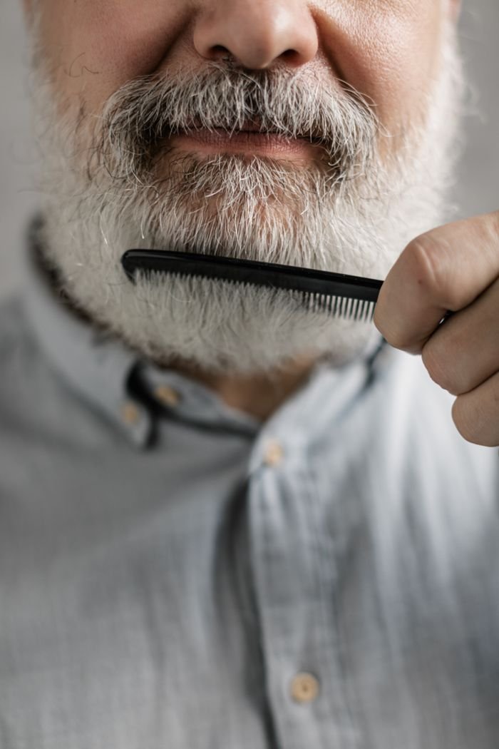 An elderly man combing his grey beard, focused on grooming and personal care.