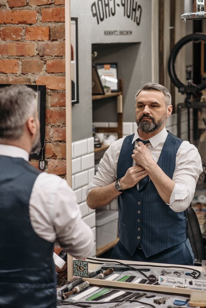 Stylish adult man adjusts his tie while looking in a barbershop mirror, showcasing modern fashion.